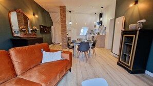 A modern living room with a brown sofa, a dining table with chairs, and a kitchen area in the background. - Appartement Tout Neuf, Tout Cocooning et Tout Équipé Pour un Excellent Séjour (Laon)