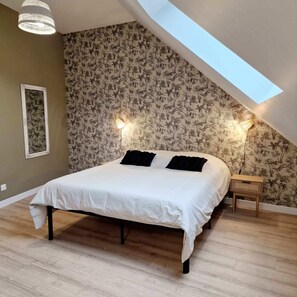 Una camera da letto con soffitto spiovente, un letto con lenzuola bianche, due cuscini neri, un comodino con una lampada e carta da parati con motivi floreali.