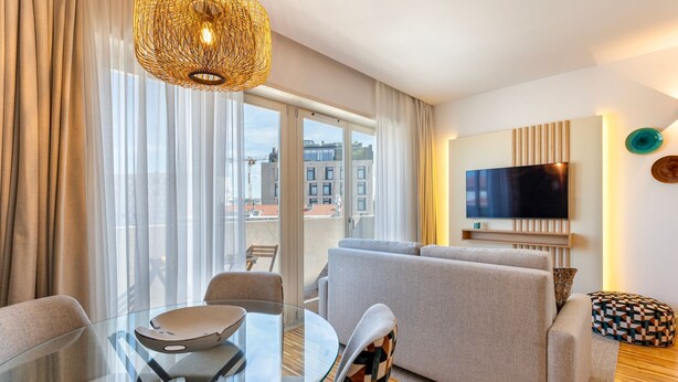 Ein modernes Wohnzimmer mit Glastisch, einer Couch, einem an der Wand befestigten Fernseher und einem Balkon mit Blick auf die Stadtlandschaft.