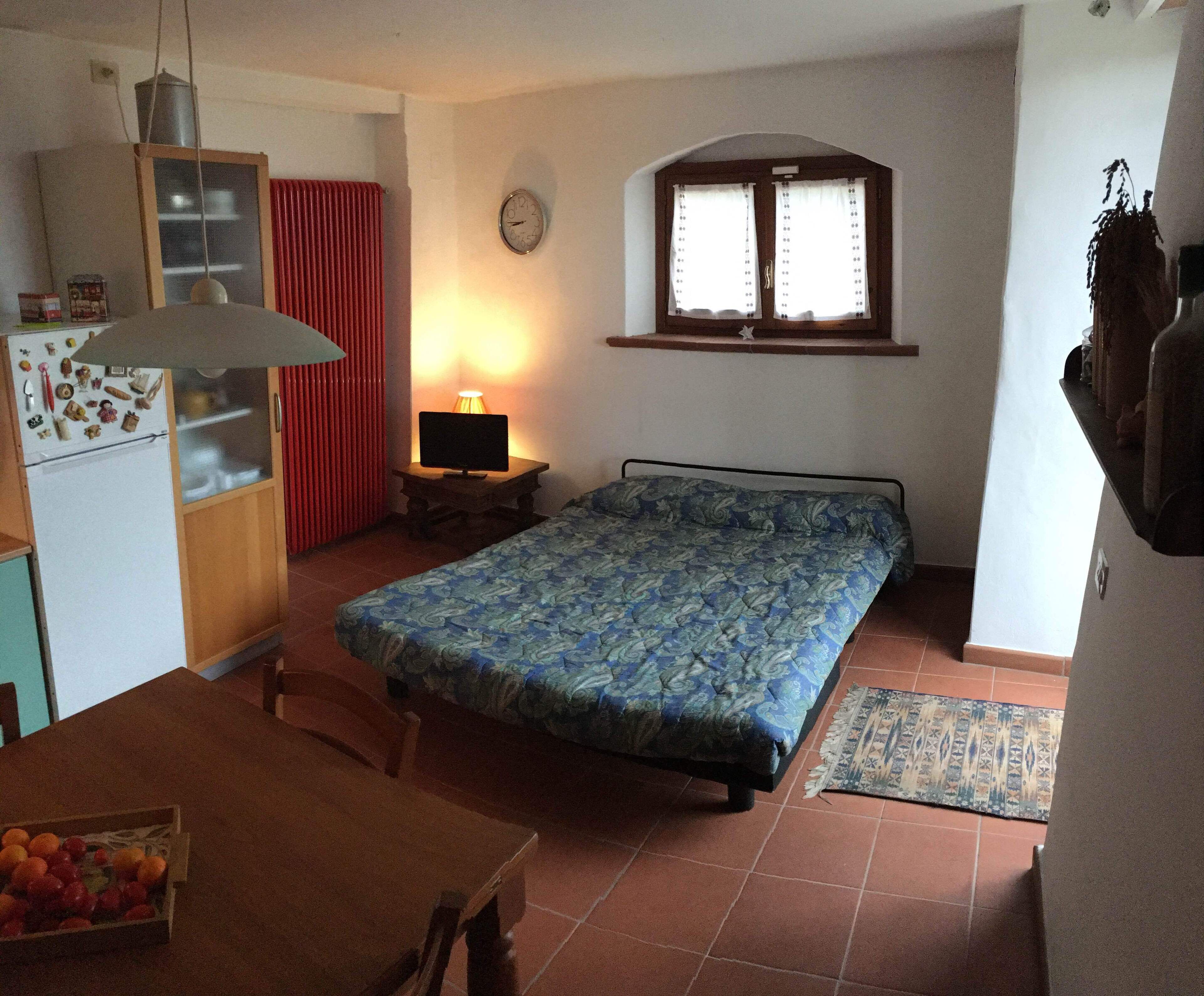 Une chambre à coucher de taille modeste, comprenant un lit, un petit bureau avec une lampe, un réfrigérateur et une fenêtre avec des rideaux.