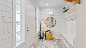 A hallway with white paneled walls, a patterned rug, a round mirror, a lamp, a wicker basket, and a shelf with hooks. - Luxury beachfront home with ocean views, pool, EV charger, chef's kitchen & golf (Port Aransas)