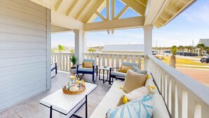 A covered outdoor seating area with a white ceiling, a white sofa with cushions, a table with a basket and drinks, and a view of the street and buildings in the background. - Luxury beachfront home with ocean views, pool, EV charger, chef's kitchen & golf (Port Aransas)