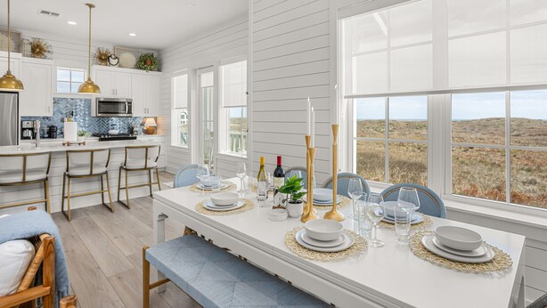 A modern kitchen with white cabinetry, a dining area with a white table set for four, and large windows offering a view of the landscape. - Luxury beachfront home with ocean views, pool, EV charger, chef's kitchen & golf (Port Aransas)