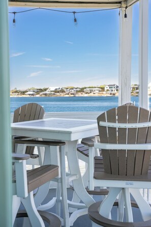 A covered outdoor seating area with a table and chairs, overlooking a body of water and a distant shoreline with houses. - Family Friendly Bayside Condo With Pool And Fishing Pier  (Pensacola)