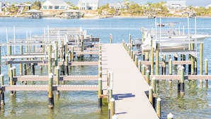A wooden pier extending into calm water with docks and boats, surrounded by houses and greenery.