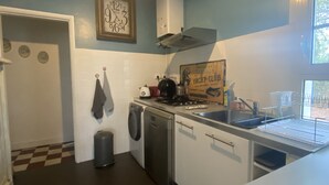 A compact kitchen with white cabinets, a stove, a sink, and a trash can. There is a clock on the wall and a sign advertising a yacht club. - Quiet house 200 meters from the ocean (Vendays-Montalivet)