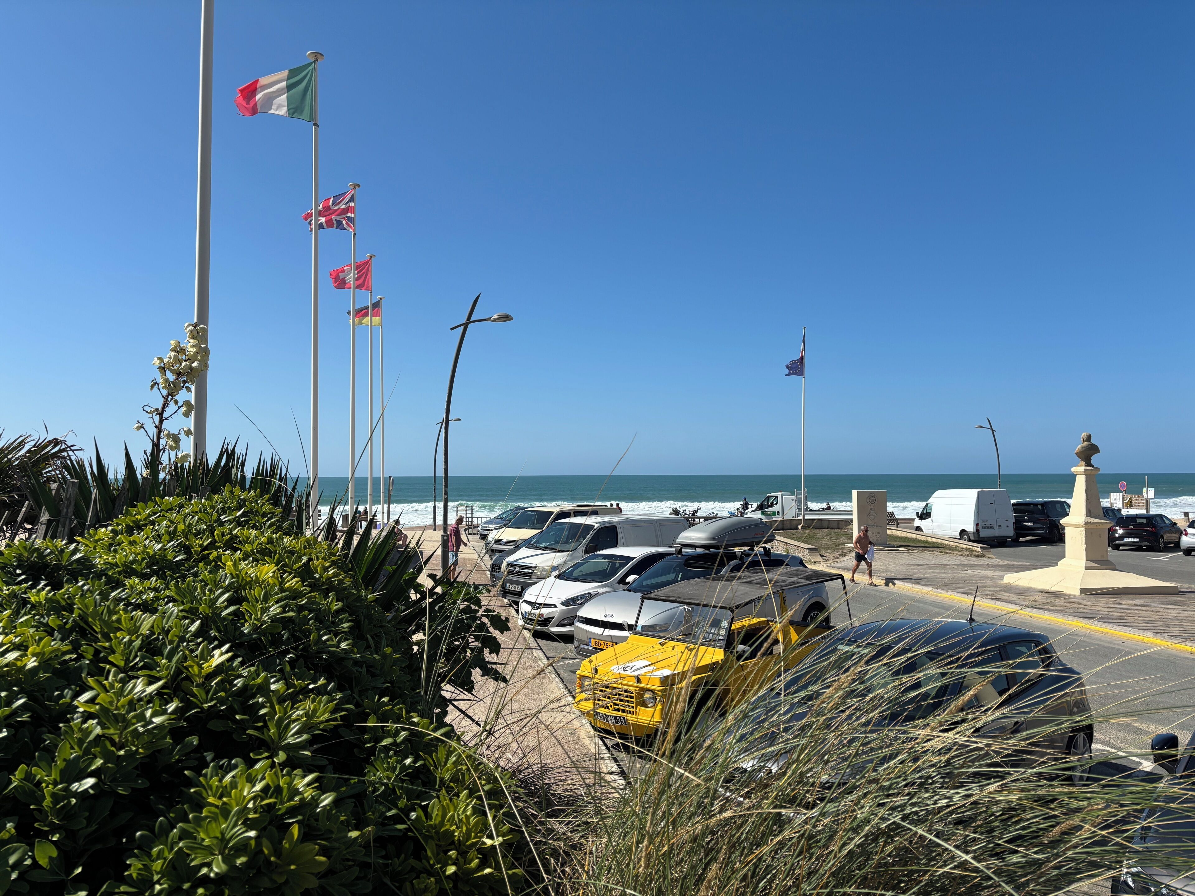 A coastal parking area with multiple flags, a clear view of the sea, and a variety of vehicles including a yellow taxi.