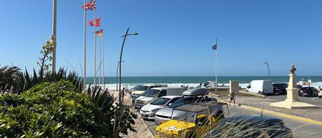 A coastal parking area with multiple flags, a clear view of the sea, and a variety of vehicles including a yellow taxi.