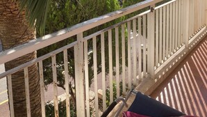 Ein Balkon mit weißer Reling, einer Palme und Blick auf einen Parkplatz mit Autos.