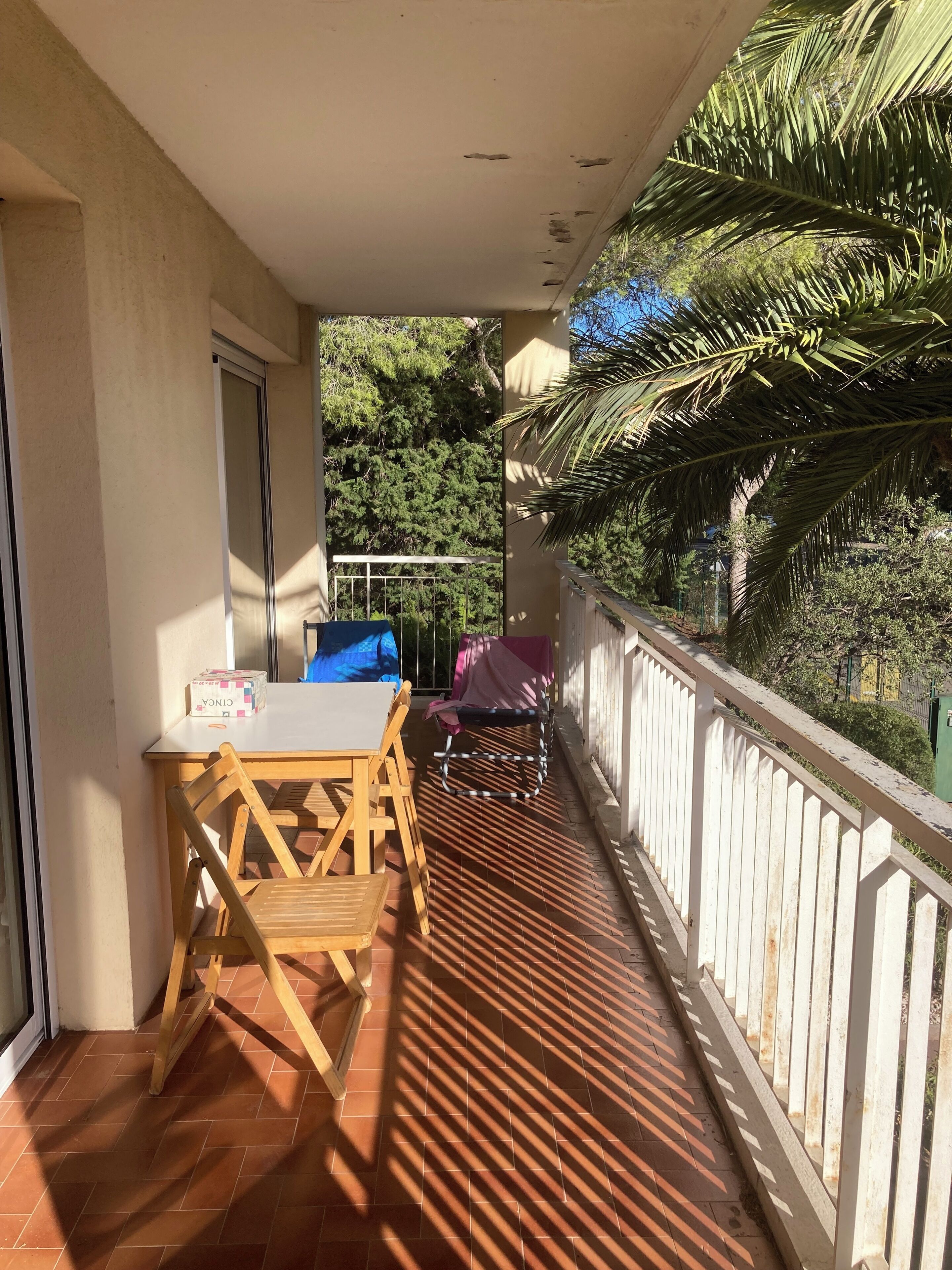 A balcony with a table and chairs, a chair with a pink cover, and a view of trees and a fence.