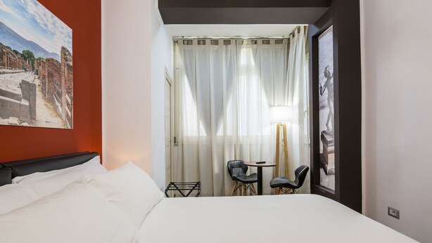 Ein Hotelzimmer mit Bett, einem kleinen Tisch mit Stühlen, einem Fenster mit Vorhängen und einem Bild an der Wand.