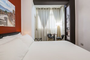 A hotel room with a bed, a small table with chairs, a window with curtains, and a painting on the wall.