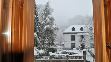 Vista a través de una puerta de madera que muestra una casa con techo nevado y árboles circundantes.