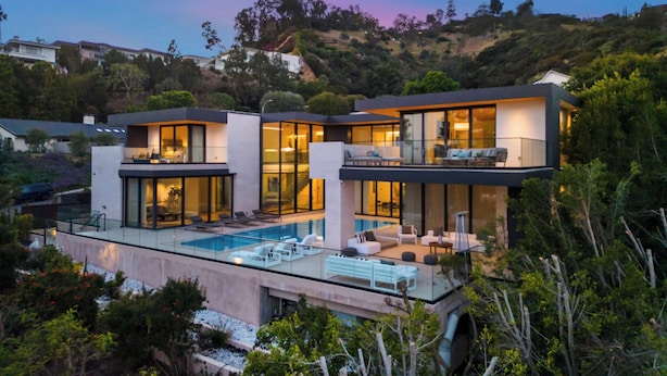 A modern two-story house with a swimming pool, large windows, and a view of a hilly landscape.
