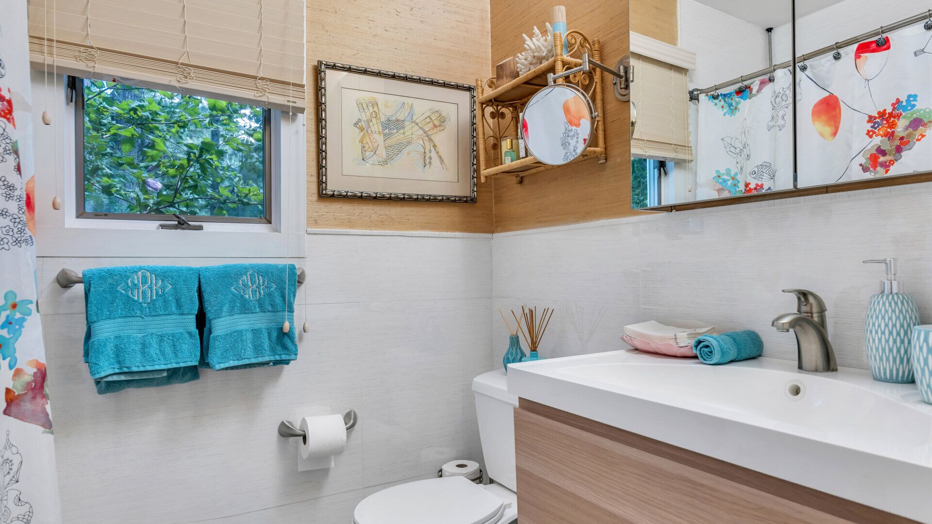 A bathroom with a toilet, sink, and window. There are towels hanging on the wall and a framed picture above the mirror.
