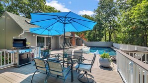 A deck with a grill, a table with chairs, a pool with swan floats, and a blue umbrella. - Beautiful East Hampton Home, Expansive Deck, Private Pool, Indoor/Outdoor... (East Hampton)