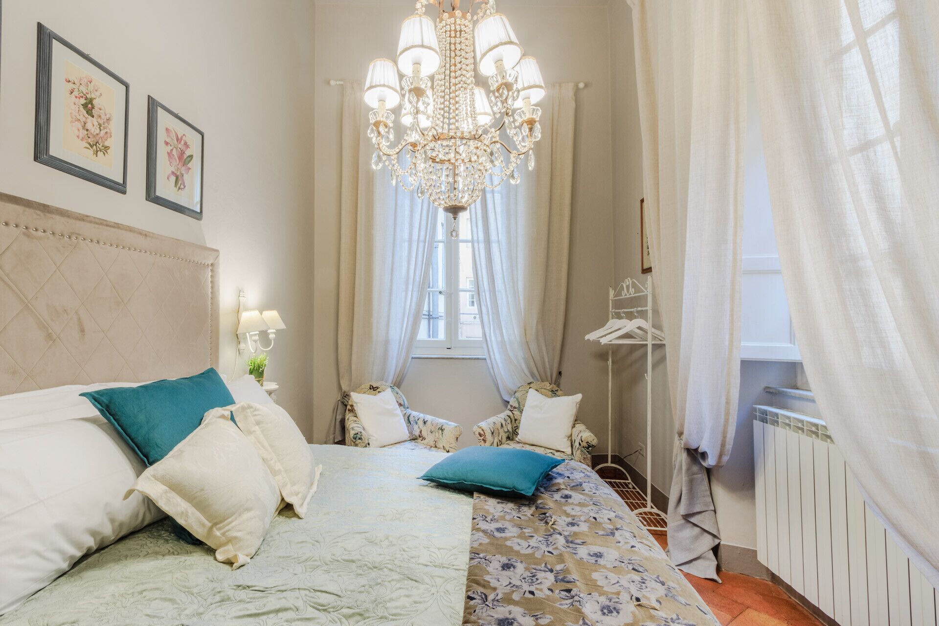 A bedroom with a bed, a chandelier, a window with curtains, and a radiator.
