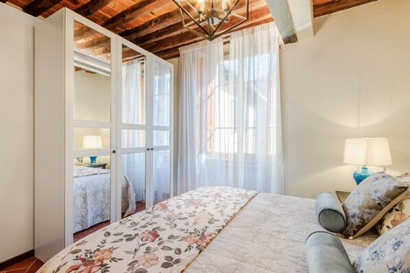 A bedroom with a bed, a nightstand, a lamp, and a window with sheer curtains.