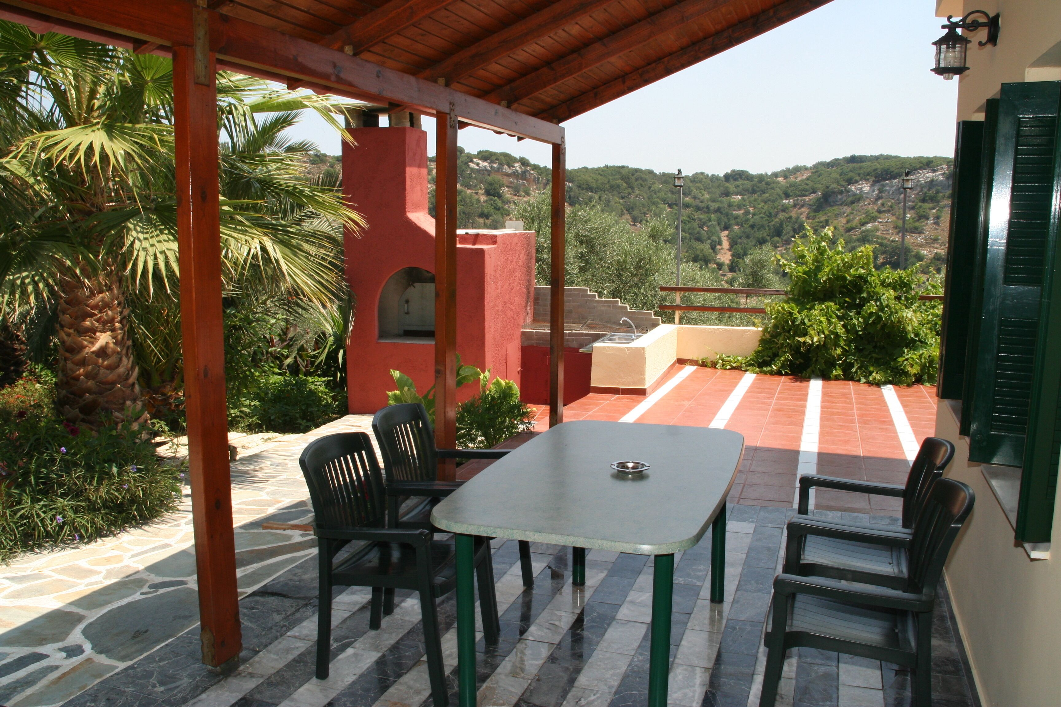 Een overdekt terras met een tafel en stoelen, een stenen vloer en uitzicht op een heuvel.