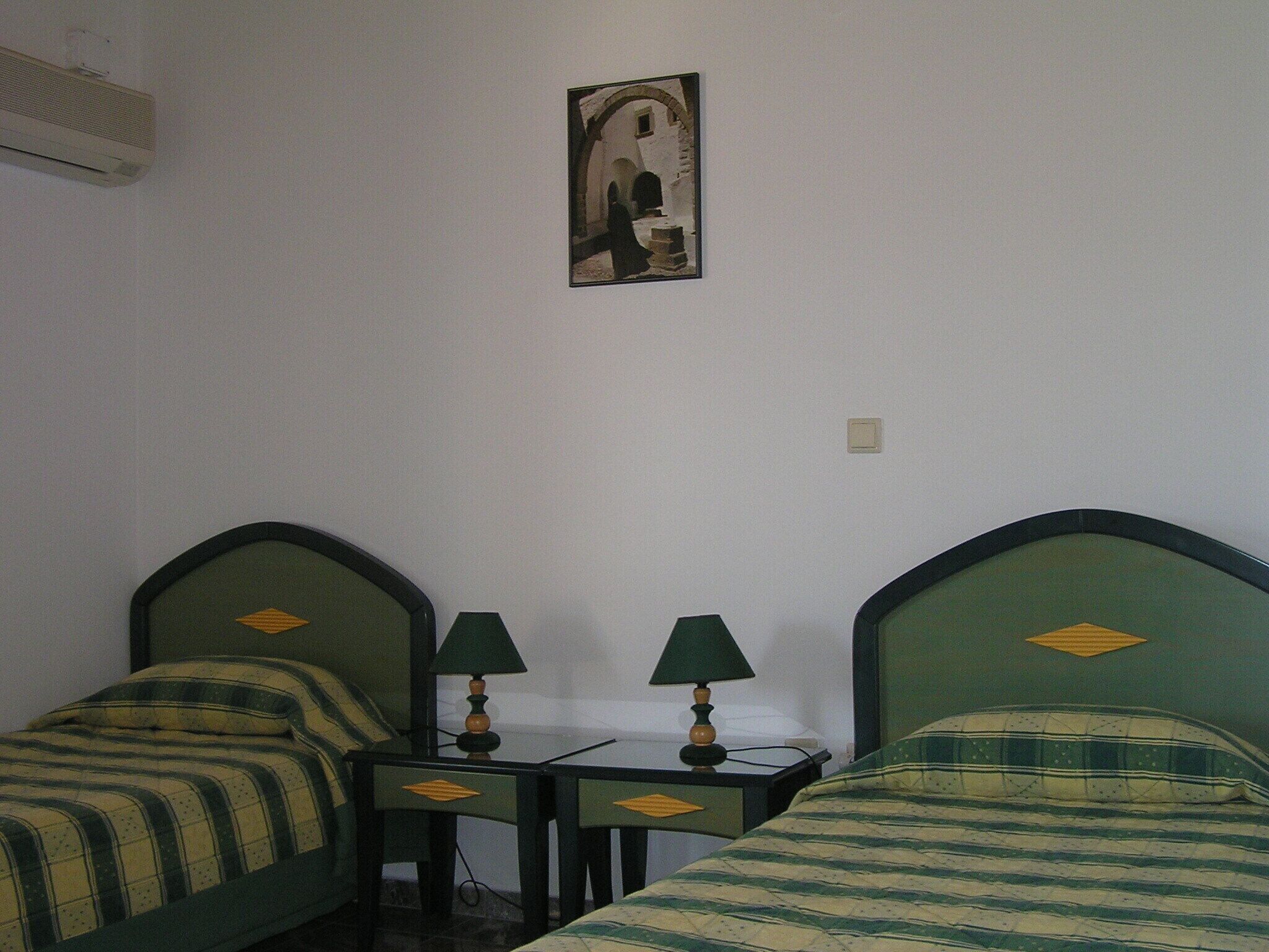 A room with two beds, each with a headboard and a pillow. There is a small table with two lamps between the beds.