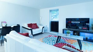 Ein modernes Wohnzimmer mit einem weißen Sofa, roten Kissen, einem Essbereich mit Tisch und Stühlen sowie einem großen Fernseher an der Wand.