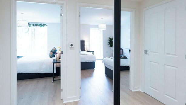 Ein modernes Hotelzimmer mit einem großen Bett, einem Schreibtisch mit Stuhl und einem kleinen Tisch mit einer Vase. Eine Tür führt in einen weiteren Raum.