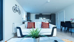 Ein modernes Wohnzimmer mit einem weißen Sofa, gestreiften Kissen, einem Topf mit Farn und einer Fernbedienung auf einem gemusterten Teppich.