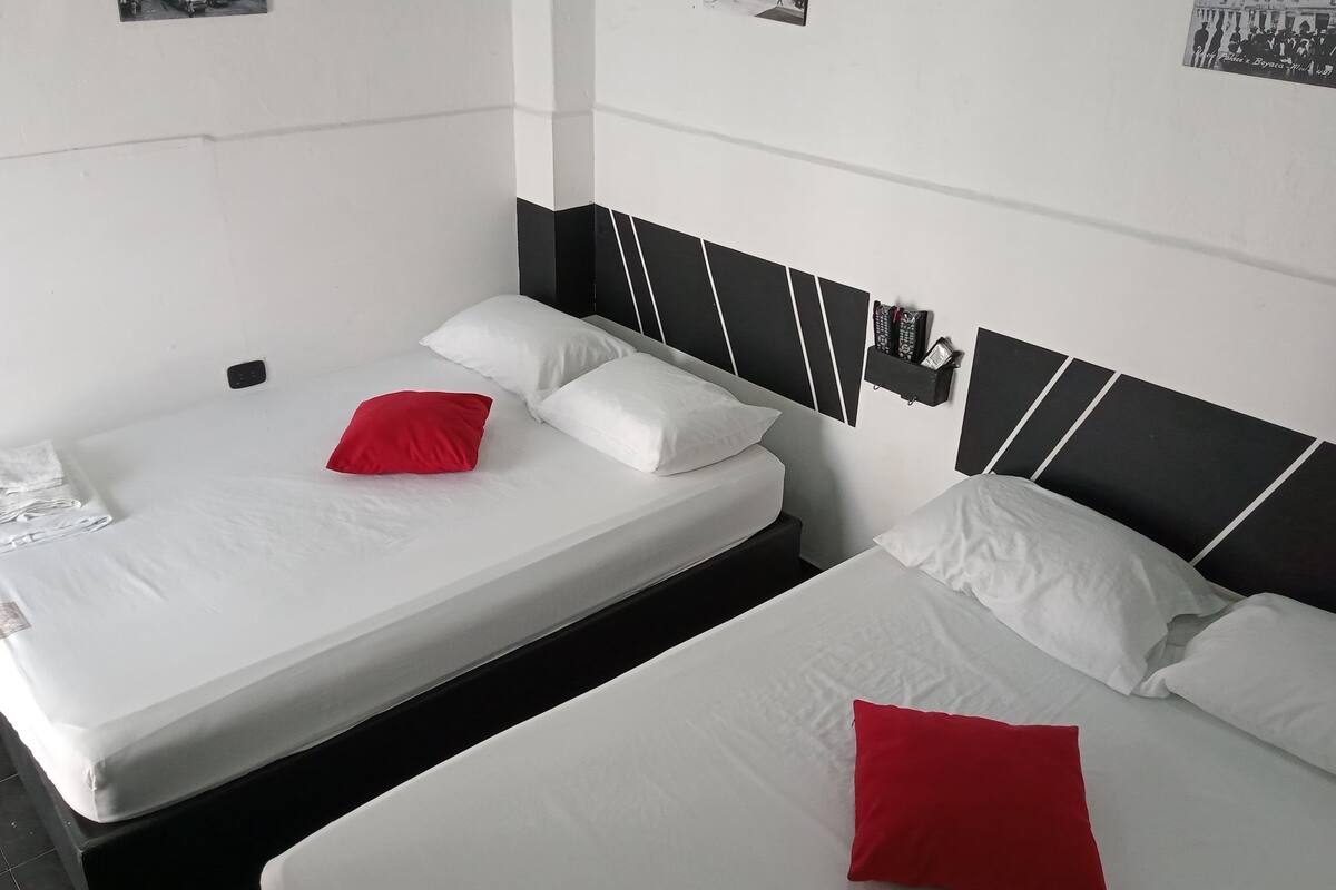 Habitación de hotel con dos camas, cada una con ropa blanca y almohadas rojas. Tiene un cabecero blanco y negro, y un estante con un control remoto y revistas.