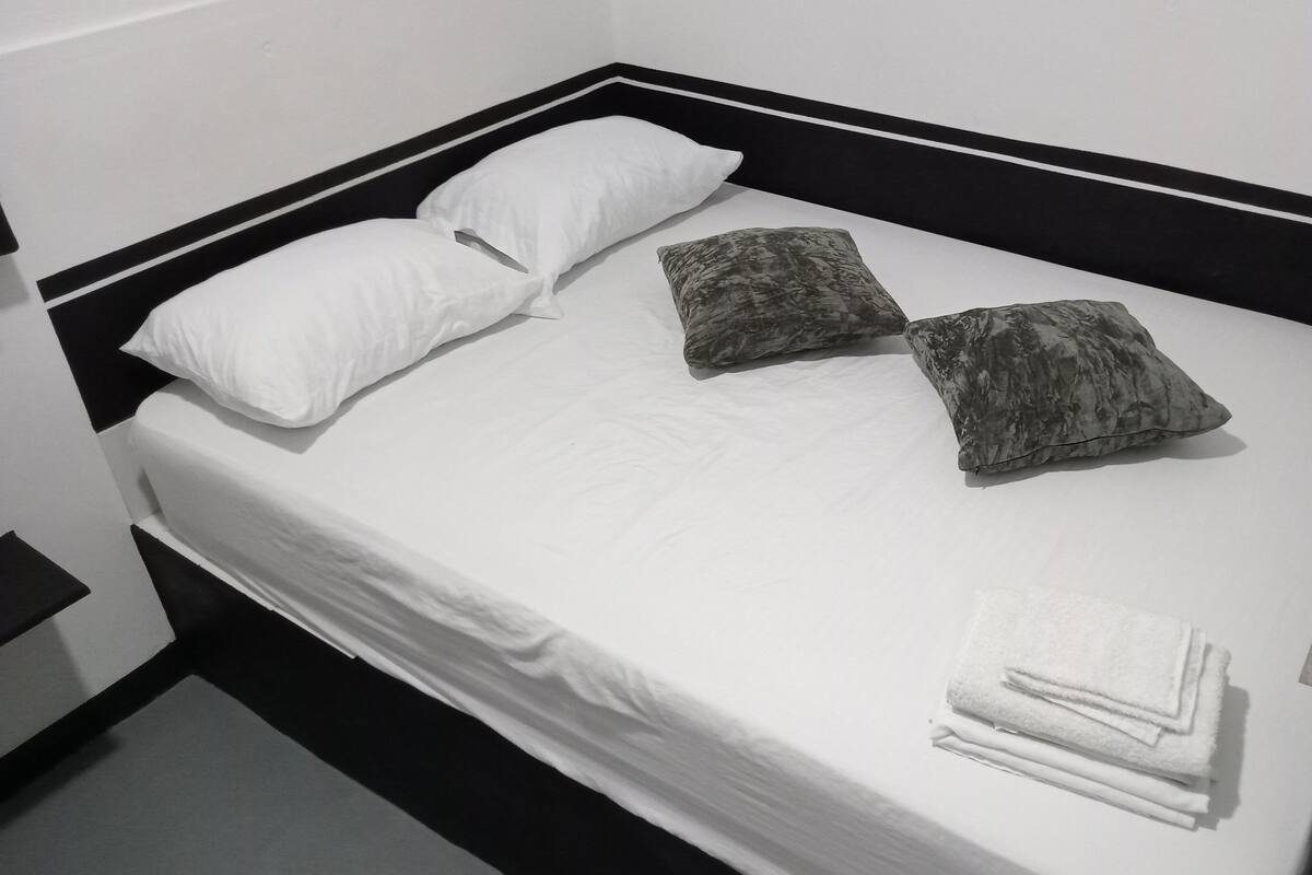 Una cama con ropa de cama blanca, dos almohadas blancas y dos almohadas grises estampadas. Al pie de la cama hay toallas blancas dobladas.