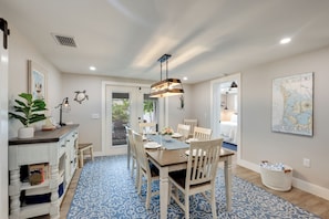 A dining area with a wooden table, chairs, a hanging light fixture, a map on the wall, and a patterned rug.