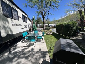 Um camping com mesa, cadeiras, churrasqueira e uma área coberta com uma árvore.