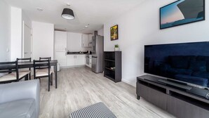 A modern living room with a flat-screen TV, a dining area with a table and chairs, and a kitchen with white cabinets and a refrigerator. - Riverside Retreat Modern Flat in Brentford (London)