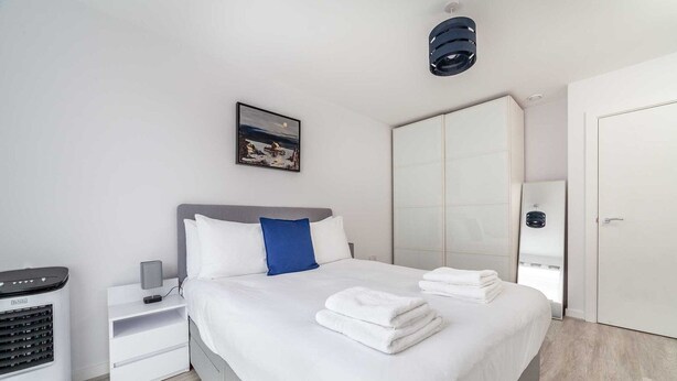 A modern hotel room with a neatly made bed, a bedside table with a lamp, a heater, and a large wardrobe. - Riverside Retreat Modern Flat in Brentford (London)