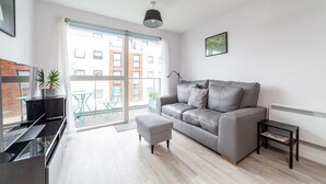 A modern living room with a grey sofa, a small table, a floor lamp, and a view of the city through large windows. - Riverside Retreat Modern Flat in Brentford (London)