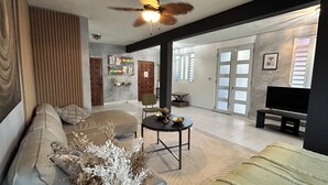 A spacious living room with a sofa, a coffee table, and a TV. There is a ceiling fan and a door leading to another room.