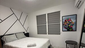 A bedroom with a bed, a window, an air conditioning unit, and a painting on the wall.