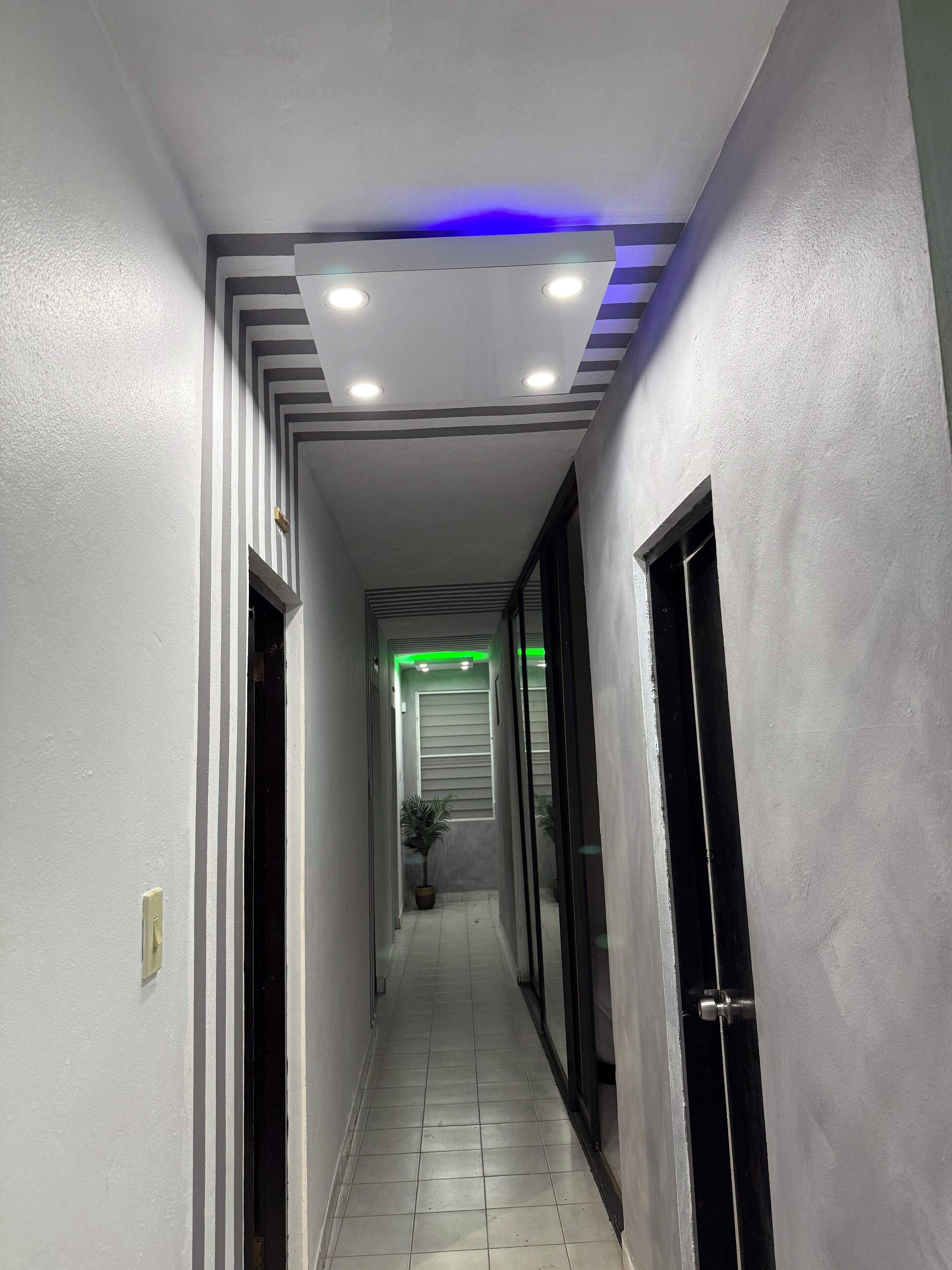 Un pasillo estrecho con paredes blancas, piso de baldosas e iluminación empotrada y una tira de luces LED azules.