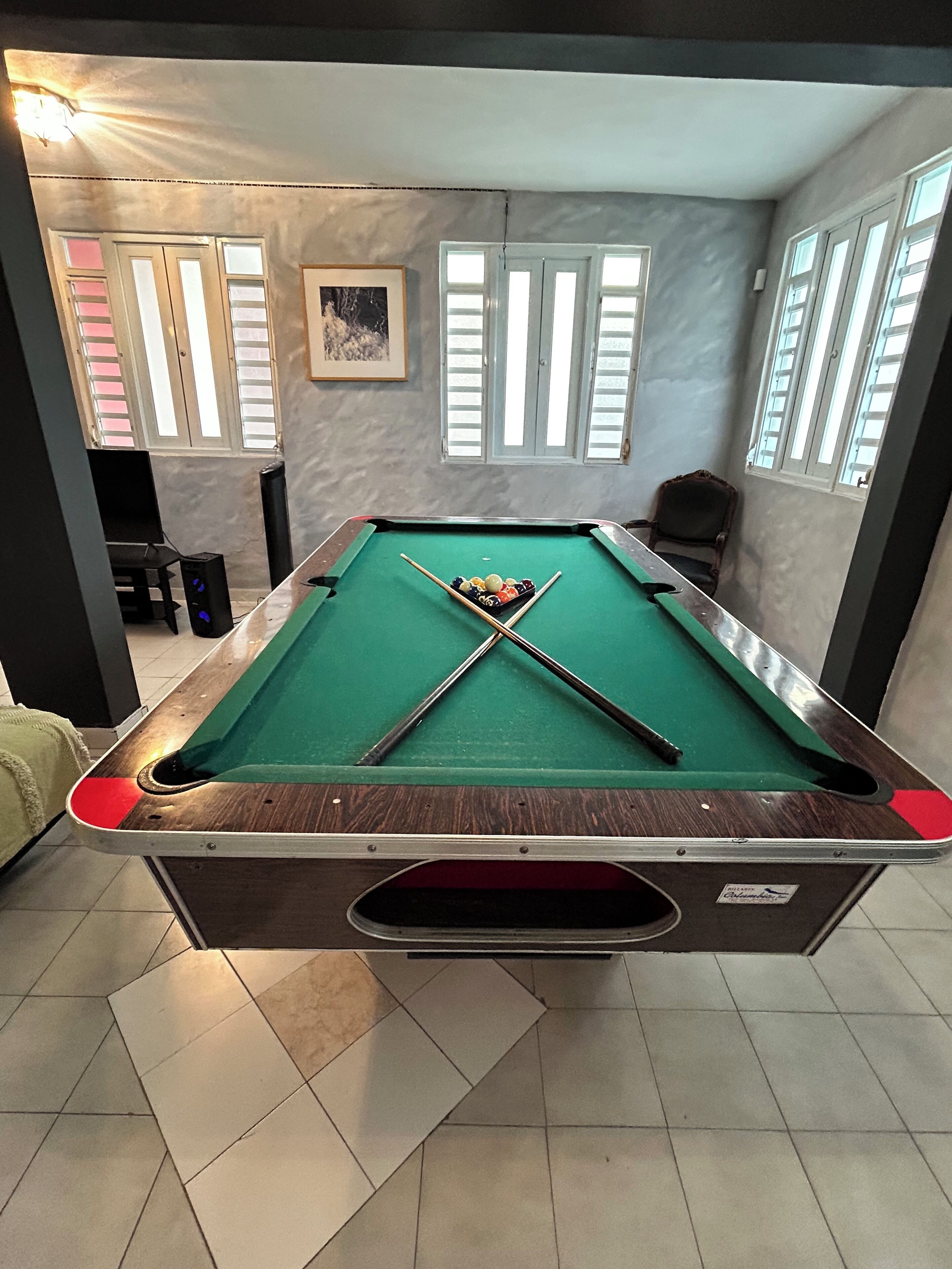 A pool table with a green felt surface, surrounded by a room with tiled flooring and large windows with blinds.