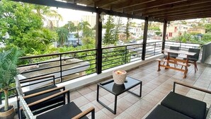 A covered outdoor seating area with modern furniture, a view of greenery and buildings, and a tiled floor.