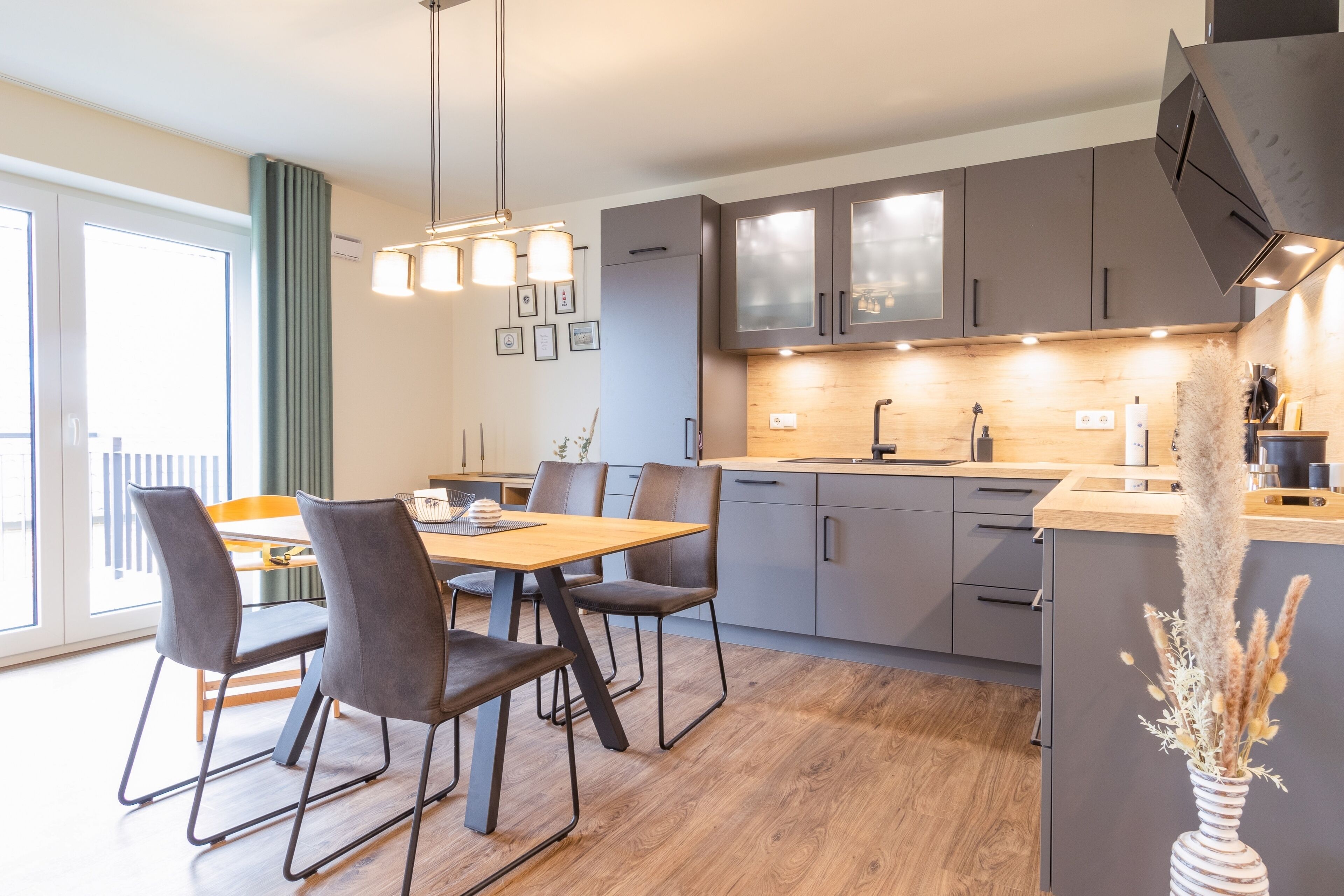 A modern kitchen with grey cabinets, a wooden dining table, and a vase with dried flowers.