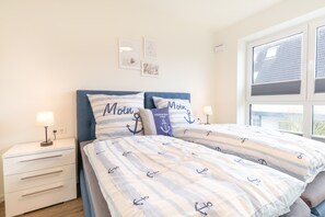 A bedroom with a bed featuring anchor-themed bedding, a bedside table with a lamp, and a window with a view of the outside.