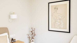 Une chambre à coucher au style minimaliste, comprenant un lit, une table de chevet, une commode, un vase avec des fleurs, une œuvre d’art encadrée et une chaise.