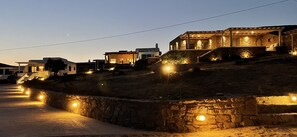 Un quartier résidentiel situé sur une colline, avec des maisons éclairées par une lumière chaleureuse, un mur de soutènement en pierre et une route pavée.