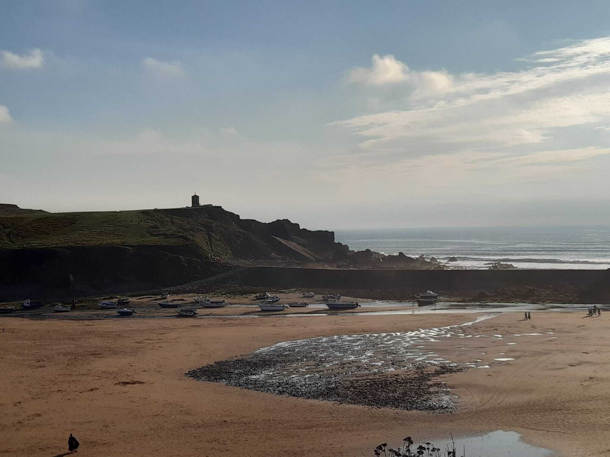 Un paisaje costero con una playa de arena, afloramientos rocosos y un faro en una colina. Barcos amarrados a la orilla.