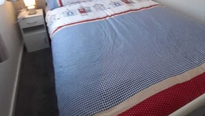 Un lit recouvert d’une couvre-lit à motifs bleus et blancs, deux oreillers décorés de motifs représentant une maison, une œuvre d’art accrochée au mur et une lampe de chevet.