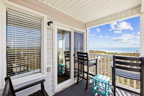 Un balcon abrité offrant une vue sur la plage, avec une table, des chaises et un petit tabouret.