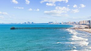 Eine Küstenstadt mit einer Pier, die ins Meer führt, einem Strand mit Menschen und einer fernen Skyline.