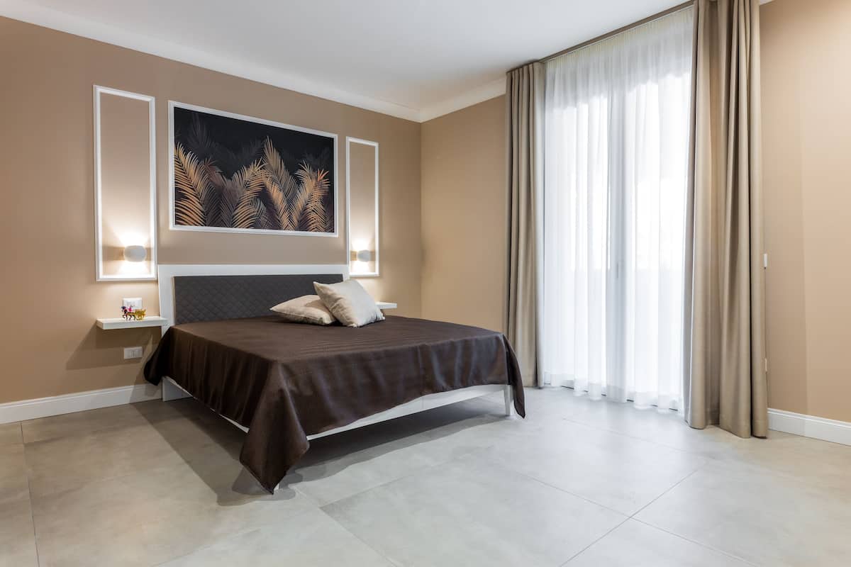 Une chambre moderne avec un grand lit, une table de chevet, une lampe fixée au mur et une grande fenêtre avec des rideaux.