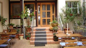 An outdoor cafe with wooden tables and chairs, potted plants, and a tiled entrance. - Hotel zum Stadttor  (Kahla)