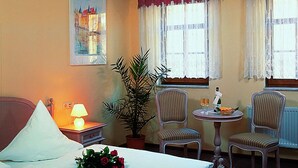 A hotel room with a bed, a nightstand with a lamp, a potted plant, two upholstered chairs, a small table with a bottle and glasses, and a window with curtains. - Hotel zum Stadttor  (Kahla)